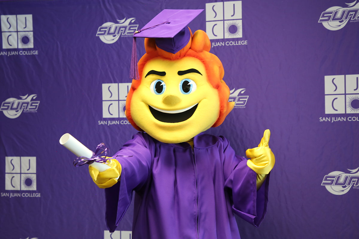 Blaze in a graduation cap and gown in front of a SJC Backdrop