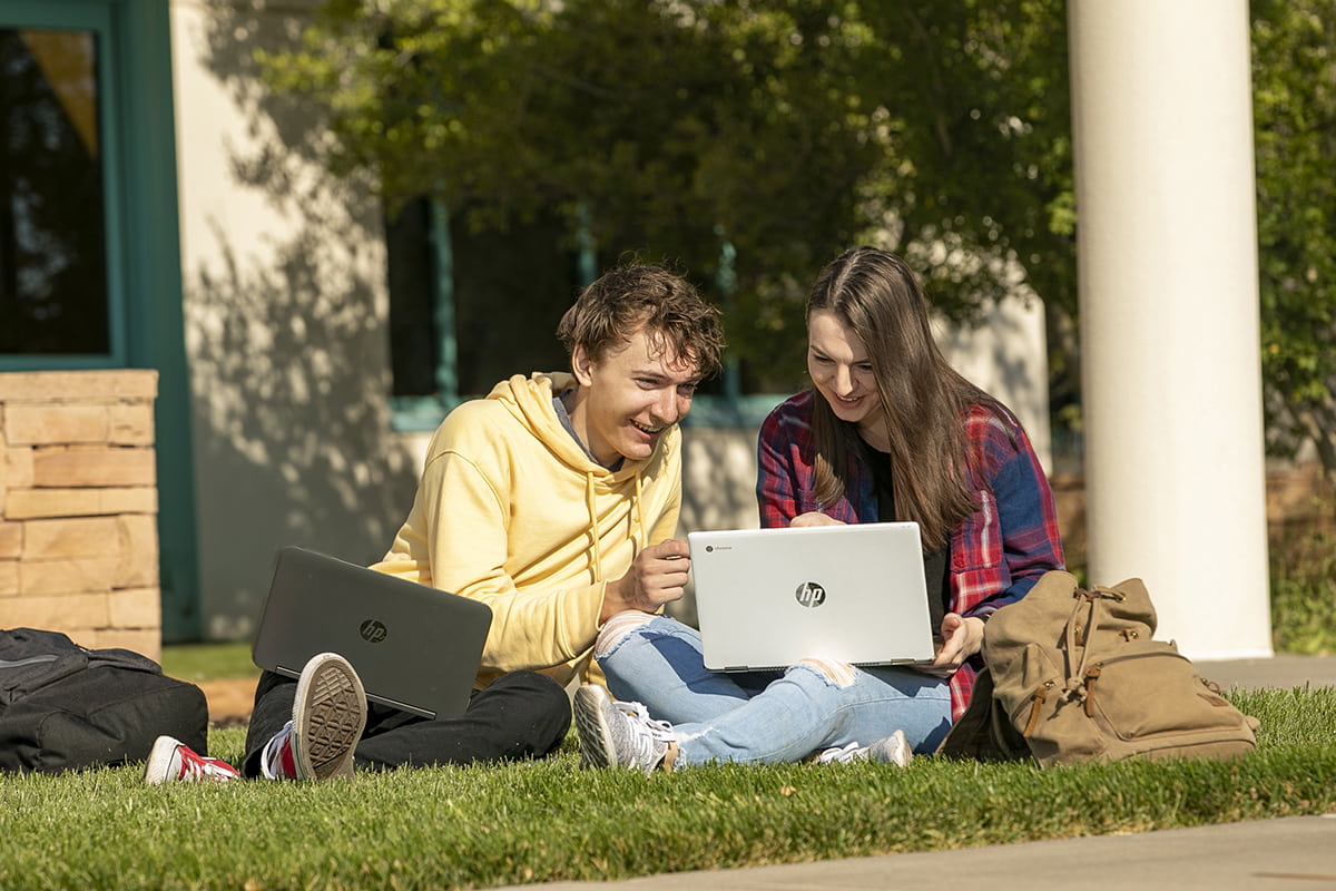 Two students sitting on the grass looking at a laptop laughing