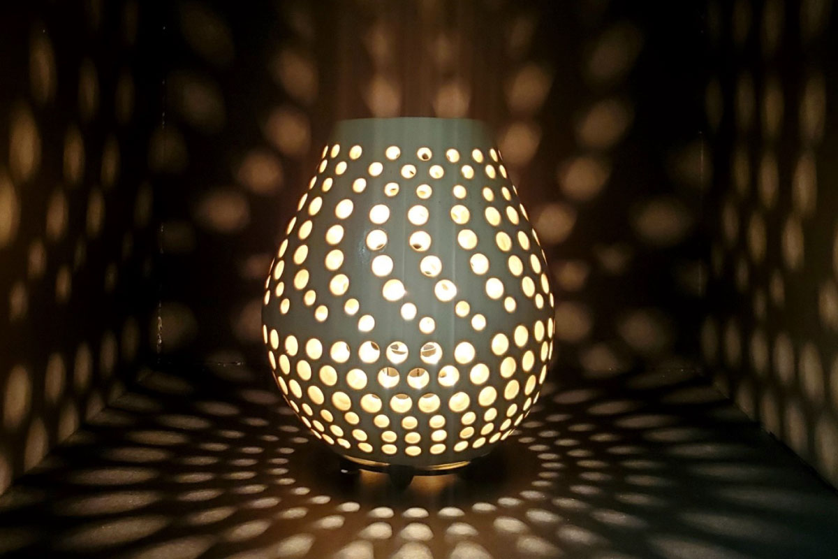 A pottery piece with holes making a spiral design with a light shining through it against a wall