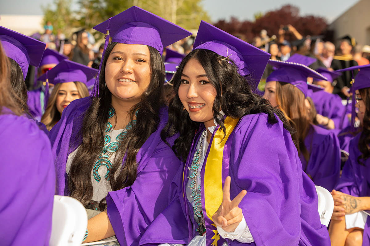 Two students in their graduation gowns. One holding up a peace sign with her left hand.
