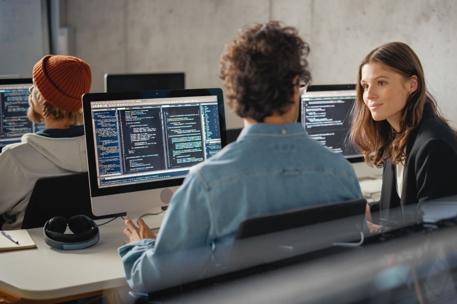 Two CompTIA students sitting in front of a computer and one of the students is talking to a CompTIA Academy instructor.