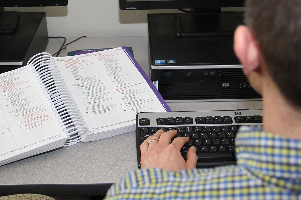 An individual sits at a computer, typing on the keyboard, with a book of medical coding information open on the desk next to them