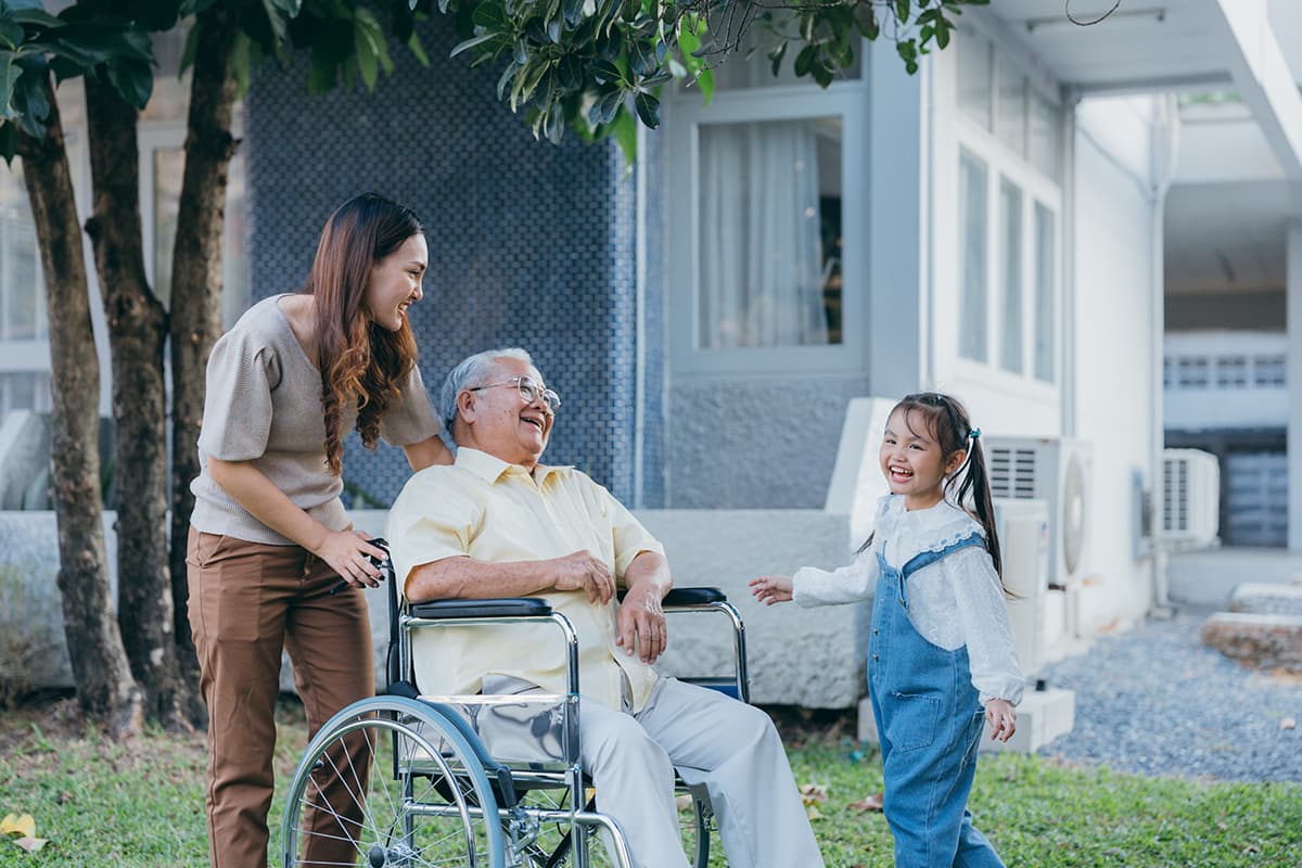 Social worker laughs along with a wheelchair bound elderly man and his grandaughter outside a house under a tree.