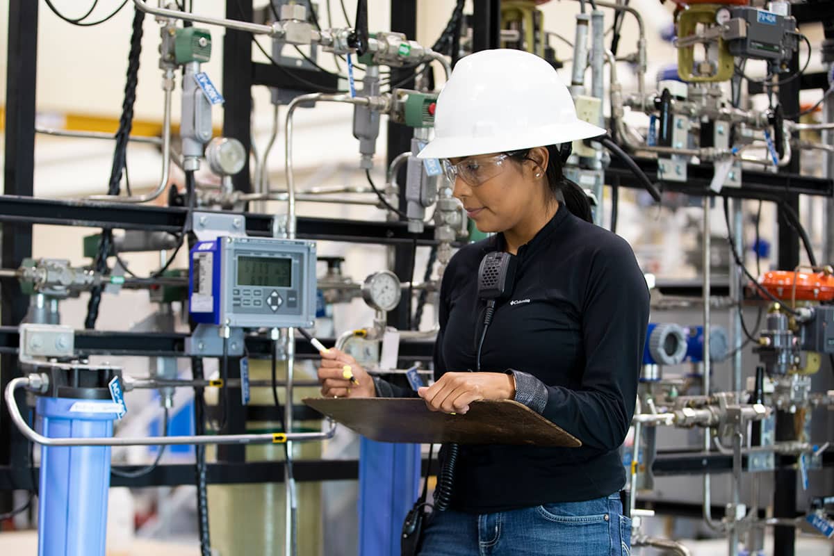 A person in a white hard hat looks at a monitor with various machines in the background