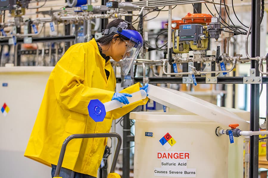 A person in a yellow lab jacket pours a beaker of solution into a container of sulfuric acid