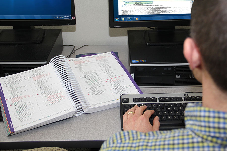 An individual sits at a computer, typing on the keyboard, with a book of medical coding information open on the desk next to them