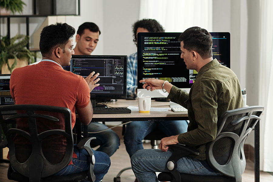 Four individuals sit in front of computers, discussing and pointing at text on the screen