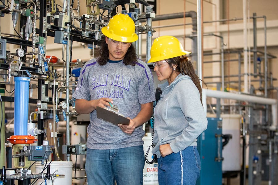 Two San Juan College students wearing hard hats checking a checklist in front of a series of pipes